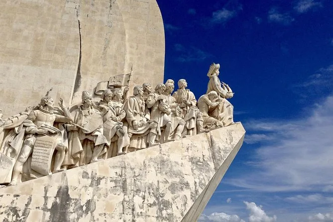 The full prow of the monument from below, the figures climbing towards Henry, under a blue sky with clouds