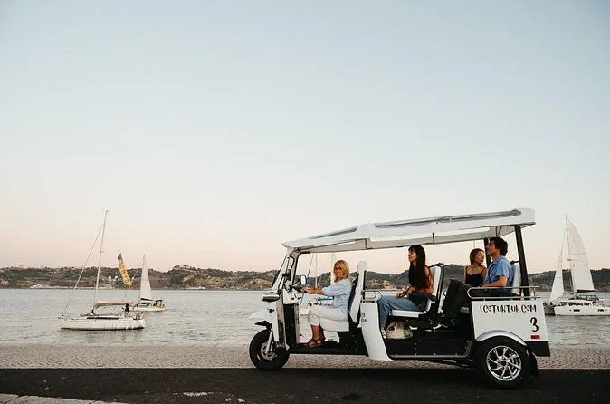 A white electric tuk-tuk parked by the river with sailboats on the water